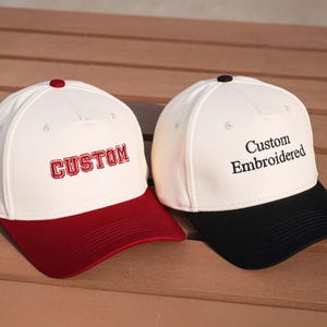 May include: Two baseball caps with white crowns and contrasting brims. One has a red brim and the word "CUSTOM" embroidered in red. The other has a black brim with "Custom Embroidered" in black. Both have a button on top.
