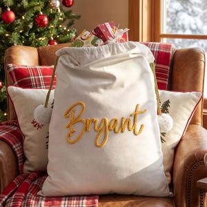 May include: A cream-colored fabric gift sack featuring the name Bryant in yellow cursive yarn lettering. The sack has a drawstring closure with gold-toned cords and white pom-poms at the ends, filled with small wrapped gift boxes.