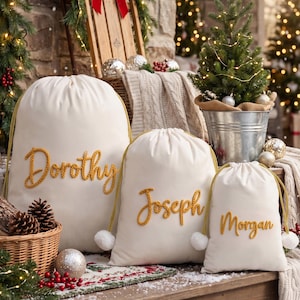 May include: Three off-white drawstring gift bags of varying sizes, each featuring a name embroidered in thick, textured gold yarn. The names Dorothy, Joseph, and Morgan are displayed on the front of the bags, which have gold cords.