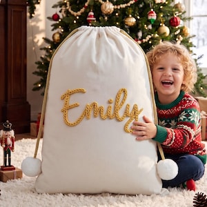 May include: A large, off-white canvas drawstring sack featuring the name Emily embroidered in thick, golden-yellow yarn. The bag includes a braided gold cord closure with two white pom-poms at the ends, providing a secure and decorative finish.