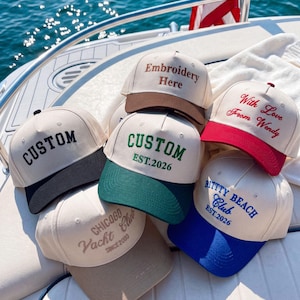 Puede incluir: Una colección de gorras de béisbol en varios colores, como beige, azul marino, verde y rojo. Cada gorra presenta texto bordado como "CUSTOM" y "Embroidery Here". Las gorras se exhiben en un barco con una bandera estadounidense al fondo.