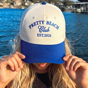 Puede incluir: Una gorra de b&eacute;isbol de color crema con una visera azul vibrante y un bot&oacute;n azul a juego en la parte superior. El frente presenta un texto azul bordado que dice Pretty Beach Club y EST. 2026 en una combinaci&oacute;n de letras de bloque y cursiva.