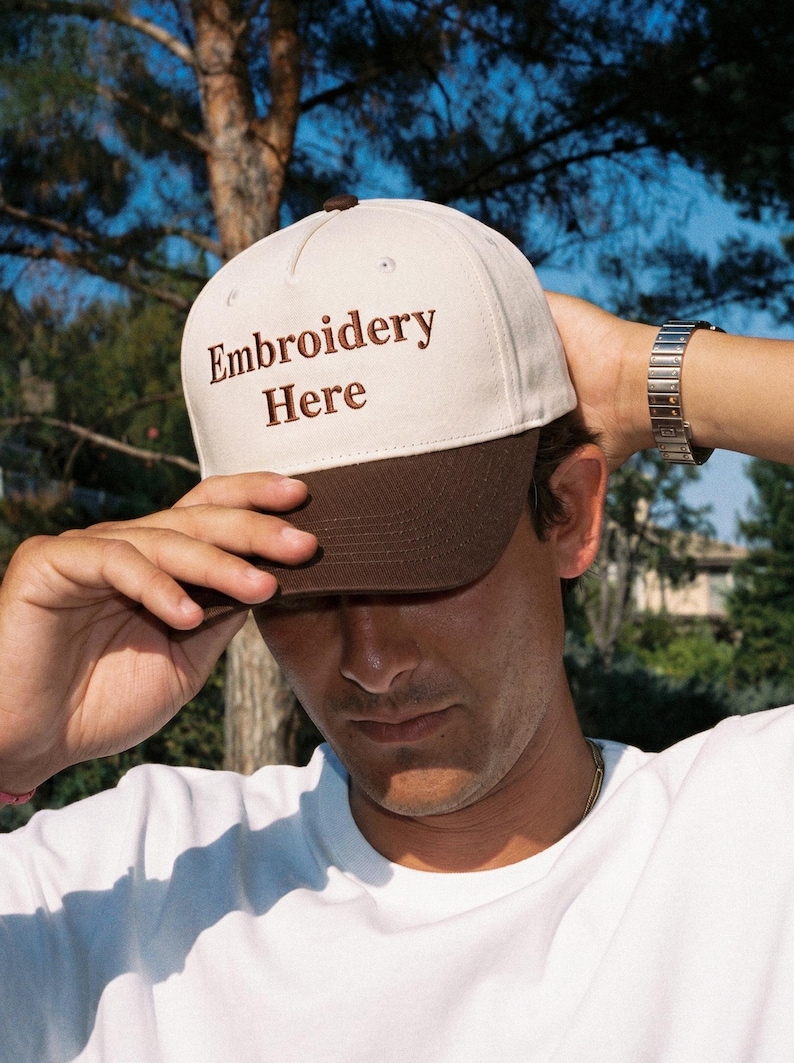 Puede incluir: Una gorra de b&eacute;isbol bicolor con una corona de color crema y una visera de color marr&oacute;n oscuro. La parte delantera de la corona muestra el texto Embroidery Here bordado en hilo marr&oacute;n oscuro. La gorra tiene un dise&ntilde;o estructurado de cinco paneles.
