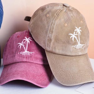 Könnte beinhalten: Drei Baseballkappen in Blau, Burgunderrot und Beige. Jede Kappe hat ein weißes, gesticktes Palmen-Design über einer wellenförmigen Linie. Die Kappen sind aus weichem, abgenutztem Stoff.