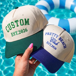 Puede incluir: Dos gorras de b&eacute;isbol con coronas de color crema. La gorra izquierda tiene una visera verde oscuro y texto bordado en verde que dice CUSTOM EST.2026. La gorra derecha tiene una visera azul y texto bordado en azul PRETTY BEACH Club EST.2026.