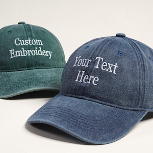 Puede incluir: Se muestran dos gorras de béisbol, una verde azulado y otra azul vaquero. La gorra verde azulado tiene bordado en blanco "Custom Embroidery". La gorra azul vaquero tiene "Your Text Here" bordado en blanco. Ambas gorras tienen visera curva.