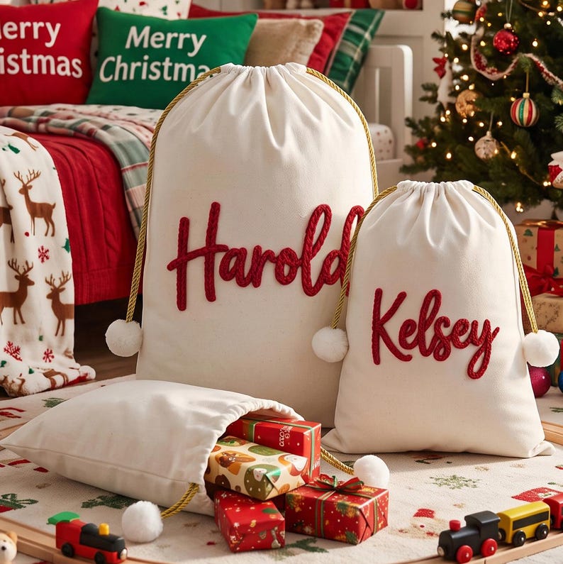 May include: Two off-white canvas drawstring bags with gold cords and white pom-poms. The larger bag features the name Harold in red script, and the smaller bag features the name Kelsey in red script. A third smaller bag lies open, revealing wrapped gifts.