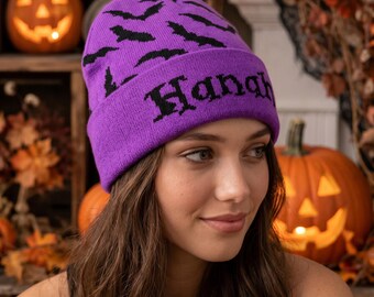 Puede incluir: Un gorro de punto de color morado brillante con un patrón repetitivo de murciélagos negros en la parte superior y la palabra "Hangry" bordada en una fuente negra de estilo gótico en el dobladillo, con un ajuste ceñido y textura.