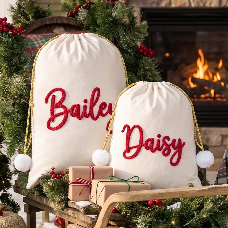 May include: Two off-white canvas drawstring bags with gold rope cords and white pom-poms. The larger bag features the name Bailey in red script, and the smaller bag features the name Daisy in red script. Both bags are positioned near wrapped gifts.
