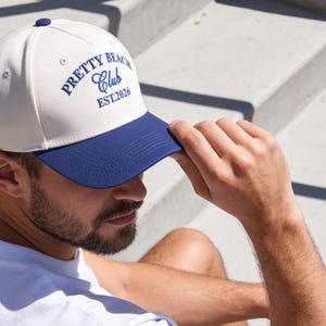 Puede incluir: Una gorra de b&eacute;isbol con corona color blanco hueso y visera azul oscuro. La parte frontal presenta un texto bordado en azul oscuro que dice PRETTY BEACH CLUB EST.2026. La gorra tiene un peque&ntilde;o bot&oacute;n azul oscuro en la parte superior.