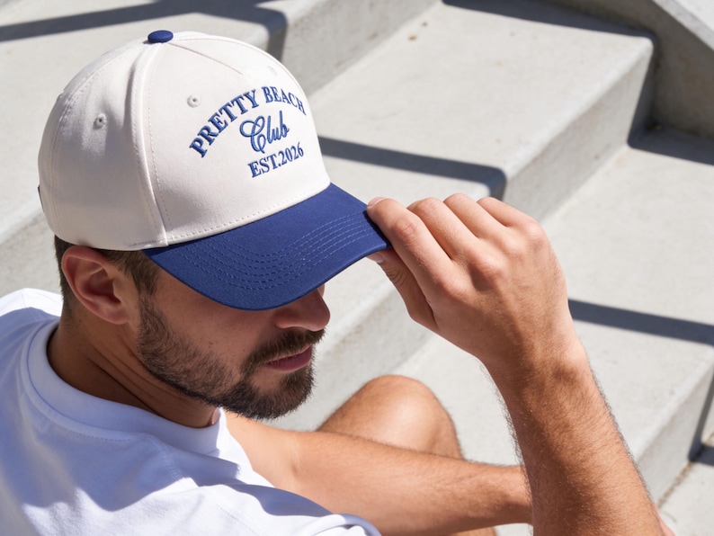 Puede incluir: Una gorra de b&eacute;isbol con corona color blanco hueso y visera azul oscuro. La parte frontal presenta un texto bordado en azul oscuro que dice PRETTY BEACH CLUB EST.2026. La gorra tiene un peque&ntilde;o bot&oacute;n azul oscuro en la parte superior.