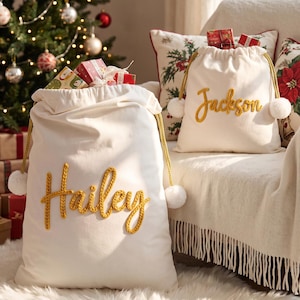 May include: Two off-white canvas drawstring gift bags featuring gold knitted lettering. One bag displays the name Hailey and the other displays the name Jackson. Each bag has gold braided drawstrings with white pom-pom ends.