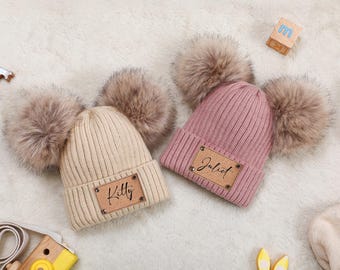 Könnte beinhalten: Zwei gestrickte Mützen mit großen Fellbommeln. Eine ist beige mit "Kitty" auf einem Lederpatch, die andere ist rosa mit "Juliet". Beide liegen auf einer weißen, flauschigen Oberfläche. Ein Holzblock und ein Spielzeug sind im Hintergrund.