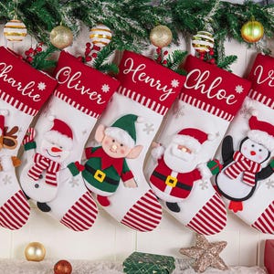 May include: Five white Christmas stockings with red cuffs featuring the names Charlie, Oliver, Henry, Chloe, and Violet. Each stocking displays a plush character: a reindeer, snowman, elf, Santa Claus, and penguin, all with red and white striped accents.