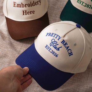 Puede incluir: Tres gorras de b&eacute;isbol con coronas de color crema y viseras en contraste. Una gorra presenta un bordado azul que dice Pretty Beach Club EST.2026, otra un bordado marr&oacute;n Embroidery Here y la tercera tiene un texto en color verde.