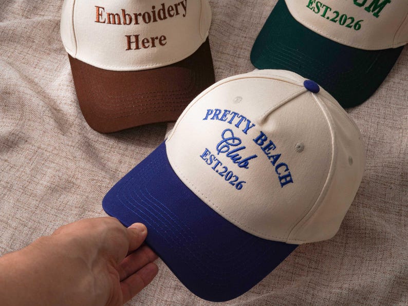 Puede incluir: Tres gorras de b&eacute;isbol con coronas de color crema y viseras en contraste. Una gorra presenta un bordado azul que dice Pretty Beach Club EST.2026, otra un bordado marr&oacute;n Embroidery Here y la tercera tiene un texto en color verde.