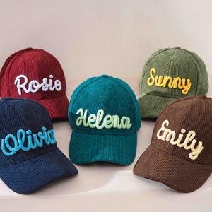 May include: Five corduroy baseball caps in various colors: navy, burgundy, teal, olive green, and brown. Each cap features a name embroidered in a contrasting color: Olivia, Rosie, Helena, Sunny, and Emily.