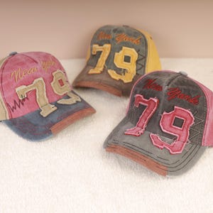 Könnte beinhalten: Drei Baseballkappen mit dem Text "New York" und der Zahl "79" im Used-Look. Die Kappen weisen eine Vielzahl von Farben auf, darunter Rosa, Grau, Beige und Gelb. Die Kappen werden auf einer weißen Oberfläche präsentiert.