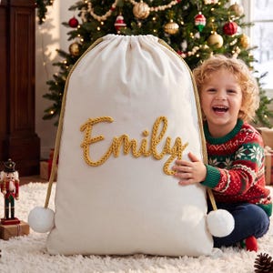 May include: A large, off-white canvas drawstring gift sack featuring the name Emily embroidered in thick, golden-yellow yarn script across the center. The bag includes gold braided rope drawstrings finished with large, fluffy white pom-poms.