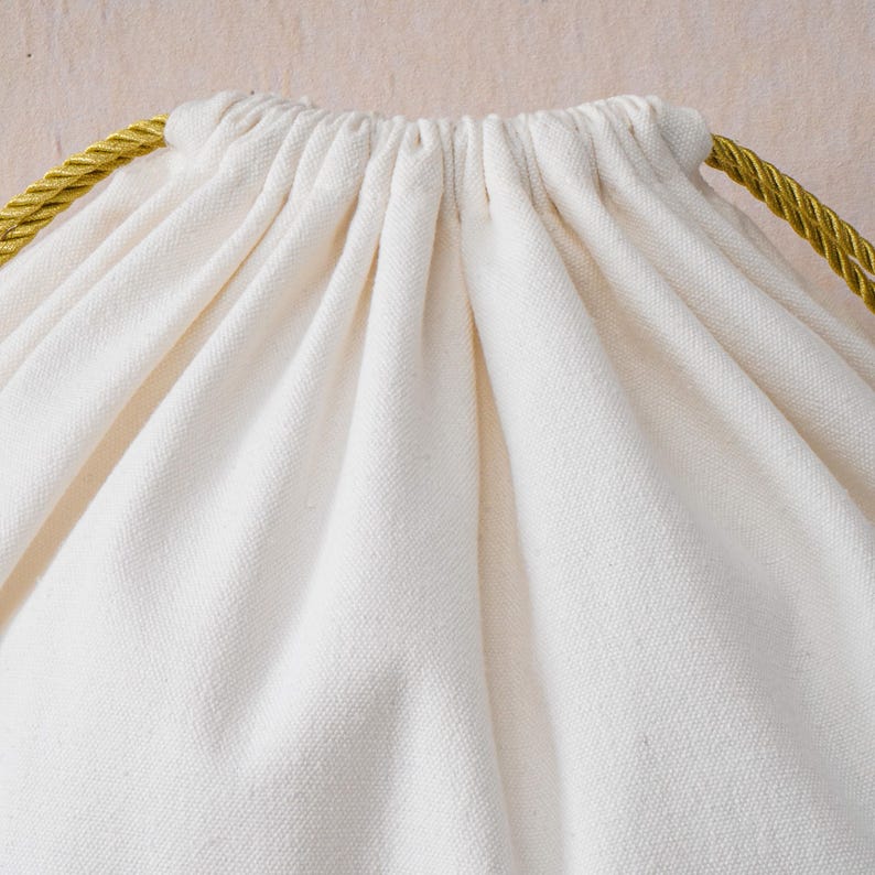 May include: A drawstring pouch made of off-white, unbleached cotton fabric. The top of the bag is gathered and cinched closed with two thick, twisted gold-colored rope cords that extend from the sides of the opening.