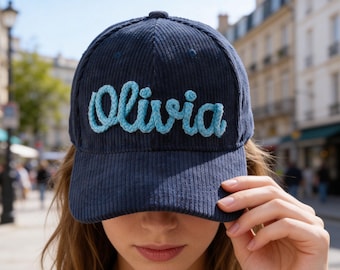 Puede incluir: Gorra de béisbol de pana azul marino con el nombre "Olivia" bordado en escritura azul claro. La gorra tiene una visera curva y un botón en la parte superior. El fondo muestra una calle de la ciudad.