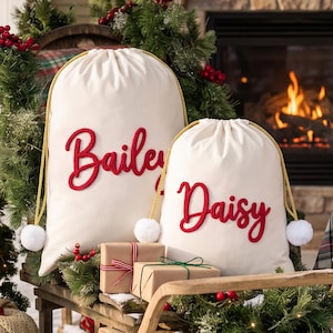 May include: Two off-white canvas drawstring bags with gold rope cords and white pom-poms. The larger bag features the name Bailey in red script, and the smaller bag features the name Daisy in red script. Both bags are positioned near wrapped gifts.