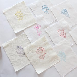 以下が含まれることがあります： 手作りの花の芸術スタンプとスタンプされた布の正方形のコレクション。スタンプは木製のブロックに取り付けられており、布の正方形には、黄色、ピンク、青などの色でさまざまな花の模様が施されています。