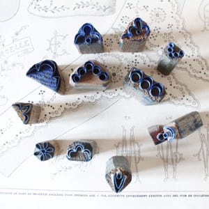 May include: A selection of antique blue and brown stamps featuring floral and geometric patterns. The stamps are displayed on a vintage paper background with text and lace designs. The stamps are made of wood and have a blue ink surface.