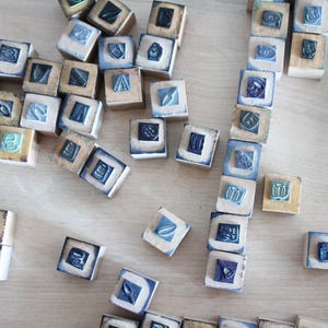 May include: A selection of small, square wooden blocks featuring embossed designs. The blocks are in natural wood tones, with blue and white detailing around the edges and within the embossed images. They are scattered on a light-coloured surface.