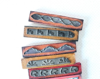Op de afbeelding: Een stapel vintage houten stempels met diverse ontwerpen. De stempels tonen patronen zoals in elkaar grijpende ovalen, gestileerde gebouwen, wolken, zonnestralen en paraplu's. Elke stempel heeft een donkergrijs ontwerp in een rechthoekig houten frame met een roodbruine rand.