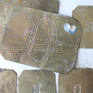Può includere: Una collezione di antiche targhe con lettere in ottone, ognuna con una lettera incisa e ornata. Le targhe hanno un aspetto ossidato e invecchiato, con segni di usura visibili. Alcune hanno piccoli fori per il montaggio. Le lettere sono in un carattere decorativo.