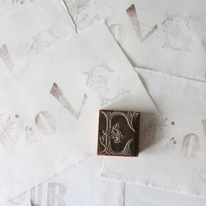 Peut inclure: Plusieurs feuilles de papier blanc cassé avec des lettres et des motifs décoratifs bruns estampillés. Un petit tampon orné d'un motif floral est au centre. Le mot "AMOUR" est partiellement visible sur une feuille, suggérant un thème d'amour ou de romance.