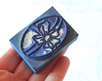 Può includere: Un timbro di legno blu con un motivo floreale. Il timbro ha una base rettangolare e un design dettagliato e in rilievo di un fiore o fiocco. Il timbro è tenuto in una mano.