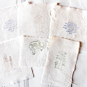 Op de afbeelding: Een verzameling kleine, gebroken witte stoffen vierkanten met handgetekende boomontwerpen in verschillende kleuren, waaronder blauw, groen en bruin. De randen van de stof zijn gerafeld, wat een rustieke uitstraling geeft. De boomillustraties variëren in stijl.