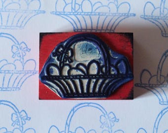 Può includere: Un timbro di gomma blu raffigurante un cesto pieno di uova, montato su un blocco di legno rosso. Il timbro viene utilizzato per creare un motivo ripetuto di illustrazioni di cesti e uova blu su sfondo bianco.
