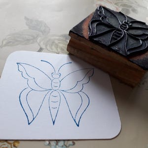 May include: A wooden block stamp featuring a butterfly design. Beside it is a white card with a blue butterfly outline. The stamp has a dark grey butterfly image on top and a wooden base.