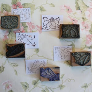 Può includere: Una collezione di timbri in legno con disegni di verdure intagliate, tra cui cavolo, peperoni e ravanelli. I timbri sono disposti con illustrazioni disegnate a mano corrispondenti su carta bianca, su sfondo floreale.