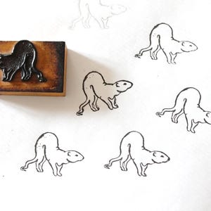 Può includere: Un timbro in legno con un disegno di ratto nero. Diversi contorni di ratto sono stampati su carta bianca. Il timbro ha una base in legno marrone scuro e una figura di ratto nero attaccata. I contorni del ratto sono in inchiostro nero.