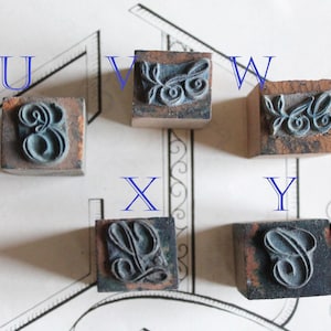 May include: Five vintage letterpress stamps, each with a different letter: U, V, W, X, and Y. The letters are in a stylized font and are made of a dark teal material. The stamps are mounted on small, square wooden blocks.