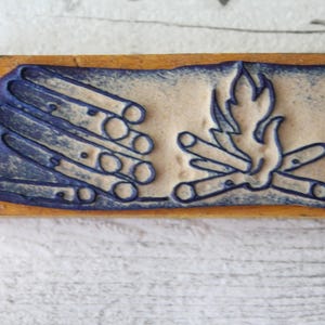 以下が含まれることがあります： 青とクリーム色のデザインの長方形の木製スタンプ。デザインは、丸太と炎のあるキャンプファイヤーが特徴です。スタンプは、工芸品や装飾要素の作成に使用される可能性があります。