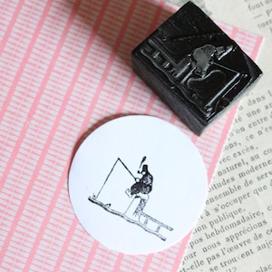 Könnte beinhalten: Ein schwarzer Stempel mit einem detaillierten Bild einer Person auf einer Leiter. Ein weißer runder Druck mit dem gleichen Bild befindet sich neben dem Stempel. Der Hintergrund ist rosa-weiß gestreiftes Papier.