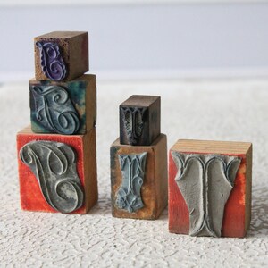 May include: A collection of vintage letterpress stamps. The stamps are made of wood and feature ornate letter designs in various colors, including gray, purple, and teal. The stamps are arranged in a stack.