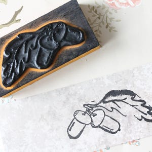 Puede incluir: Un sello de goma negro con la silueta de un perro sobre un bloque de madera, junto a una imagen estampada de bellotas y hojas. El sello tiene una veta de madera oscura y un borde amarillo. La imagen estampada es en tinta negra.
