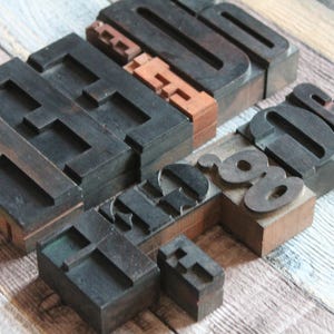 May include: A collection of vintage wooden letterpress blocks featuring various uppercase and lowercase characters. The blocks are crafted from dark brown wood with raised, ink-stained surfaces, displaying a mix of serif and sans-serif typography.