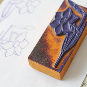 Op de afbeelding: Een rechthoekige houten stempel met een uitgesneden paars narcisontwerp. De stempel is oranje met donkerbruine randen en ligt op een wit oppervlak met een narcisschets. Voor knutsel- en kunstprojecten.