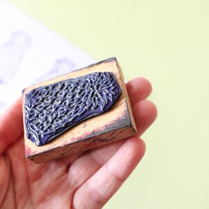 Könnte beinhalten: Ein Holzstempel mit einem dunkelblauen, strukturierten Design. Der Stempel wird in einer Hand gehalten, mit einem hellgrünen Hintergrund. Ein gestempeltes Bild ist auf einem weißen Papier im Hintergrund zu sehen.