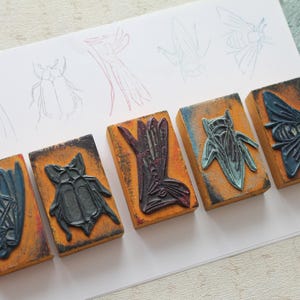 Op de afbeelding: Een set van drie keramische mokken met een gespikkelde, crèmekleurige glazuur. Elke mok heeft een ander handgeschilderd bloemmotief in tinten blauw, groen en geel. De mokken hebben een eenvoudige, afgeronde vorm en een comfortabel handvat.