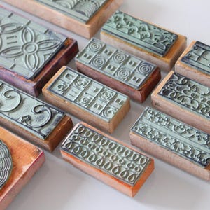 Può includere: Una collezione di timbri in gomma vintage con vari motivi decorativi. I timbri presentano disegni floreali, geometrici e astratti in un colore verde tenue, montati su blocchi di legno. I timbri sono disposti su una superficie bianca.