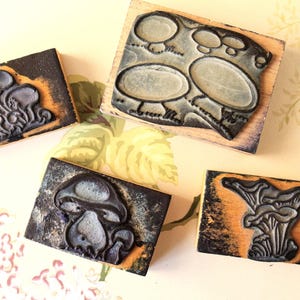 Könnte beinhalten: Vier Gummistempel mit Pilzmotiven, montiert auf Holzblöcken. Die Stempel zeigen detaillierte Illustrationen verschiedener Pilzarten in Schwarz, Grau und Orange. Sie sind auf einer Oberfläche mit floralem Muster angeordnet, ideal für Bastel- oder Kunstprojekte.