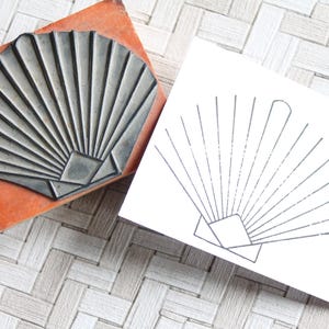 Può includere: Un timbro di gomma grigio con un disegno a ventaglio, appoggiato su un blocco arancione. Un contorno a ventaglio abbinato è stampato su una carta bianca. Il timbro e la stampa sono su una superficie strutturata dai toni neutri.